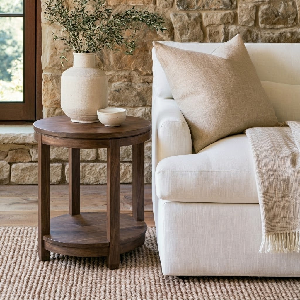 The Adams Round End Table, handcrafted from solid wood by skilled American craftsmen and finished with a durable top coat, holds a vase of greenery and a small bowl beside a white sofa accented with beige pillows and a blanket, all set against the backdrop of a stone wall and window.