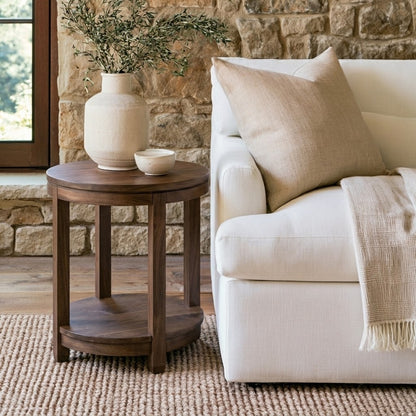The Adams Round End Table, handcrafted from solid wood by skilled American craftsmen and finished with a durable top coat, holds a vase of greenery and a small bowl beside a white sofa accented with beige pillows and a blanket, all set against the backdrop of a stone wall and window.