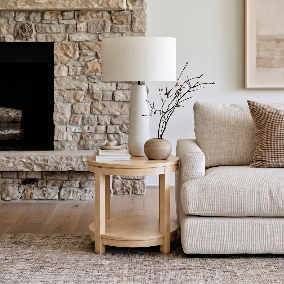 An elegant beige sofa is positioned beside The Adams Round End Table, which is expertly handcrafted from solid wood. The table is styled with a lamp, a vase with branches, and a pillow. In the background, a stone fireplace and neutral wall art enhance the space’s inviting ambiance.