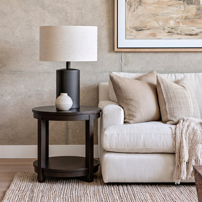 The Adams Round End Table, handcrafted from solid wood by skilled American craftsmen and finished with a durable top coat, is styled with a black lamp, a small white vase, and placed beneath a framed abstract painting above a white couch adorned with beige textured pillows and a knit throw, all set against a concrete wall.