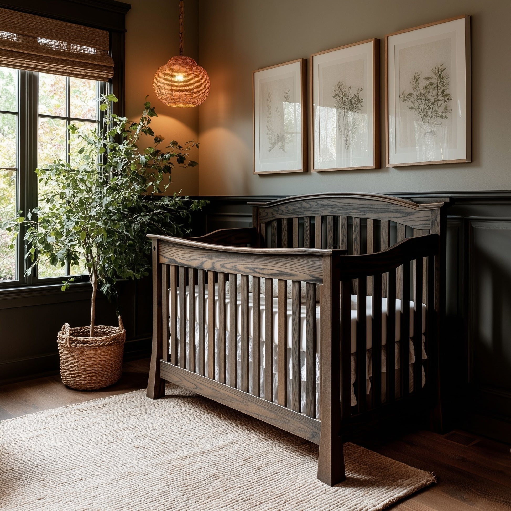 A cozy nursery with The Archer Crib in dark wood, a woven rug, a large potted plant, three botanical prints on the wall, a wicker pendant light, and sunlight streaming through window blinds.