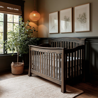 A cozy nursery with The Archer Crib in dark wood, a woven rug, a large potted plant, three botanical prints on the wall, a wicker pendant light, and sunlight streaming through window blinds.