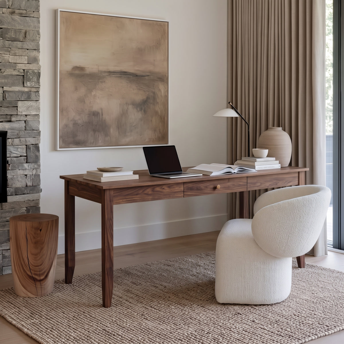 A styled home office scene featuring a dark wood writing desk with a laptop, books, and a lamp, paired with a cream boucle office chair. The setup is styled on a woven jute rug with a stone wall, curtains, and a large abstract painting in the background.