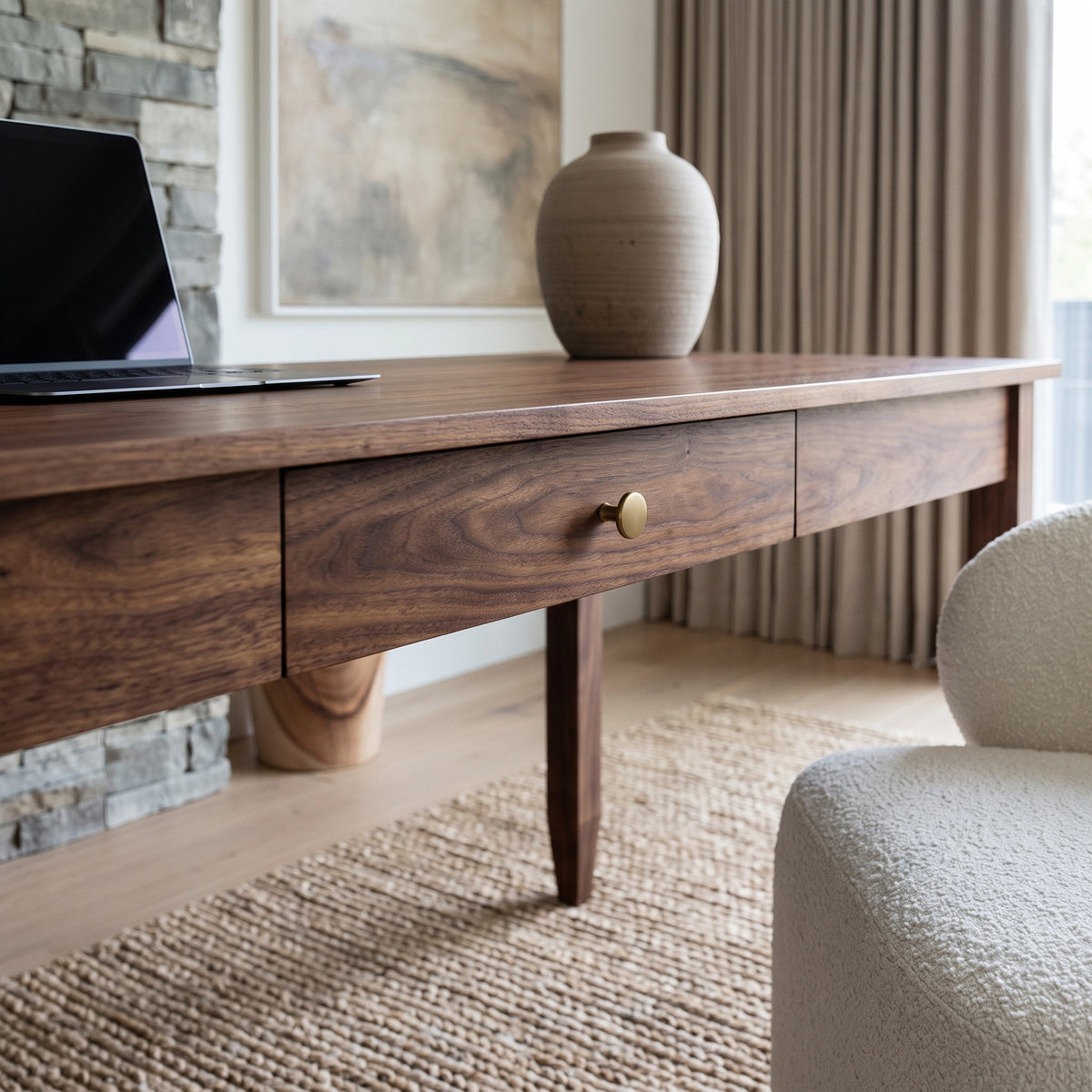 A mid-century modern wooden writing desk with a single drawer and a brass knob, styled in a bright room with a vase, laptop, and textured rug.