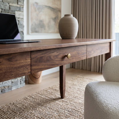 A mid-century modern wooden writing desk with a single drawer and a brass knob, styled in a bright room with a vase, laptop, and textured rug.