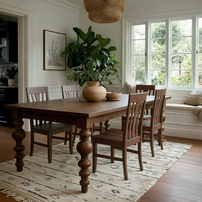 A dining room with The Aubrey Dining Table, a solid walnut design with extension leaves, six matching chairs, potted plants, ceramic vases, a patterned rug, large windows, and bench seating. Natural light warms this cozy handcrafted USA space.