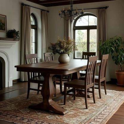 A dining room featuring The Campbell Dining Table and six chairs on a patterned rug, with a floral centerpiece, large curtained windows, a chandelier, a potted plant, and a fireplace. Handcrafted in the USA for exceptional quality.