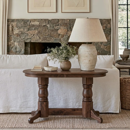 The Colt Semi-Oval Console Table, handcrafted from solid wood and finished with a durable top coat to preserve its beauty for years to come, sits adorned with decorative vases and a large lamp in front of a white sofa. Behind the arrangement, a stone fireplace, neutral artwork, and expansive windows draped with curtains contribute to the inviting, cozy atmosphere of the living room.