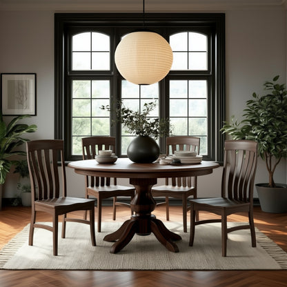 The Drayton Dining Table, a round solid walnut pedestal table with four matching chairs, sits on a light rug in a bright room. A black vase with greenery rests on the table, surrounded by large windows, plants, and framed art.