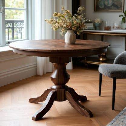 The Drayton Dining Table, crafted from solid walnut and topped with a vase of flowers, stands in a bright, elegant room with wooden floors, a window, and a gray upholstered chair nearby.