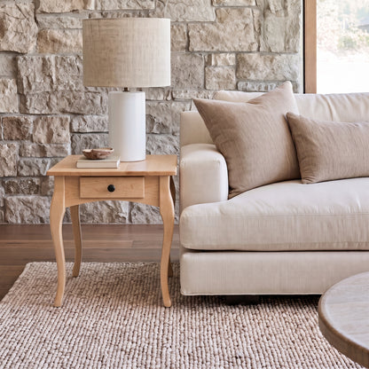 The Fox End Table, a handcrafted solid wood piece, stands beside a beige sofa with tan pillows, topped with a lamp and small bowl. All are arranged on a textured rug against a stone wall backdrop.
