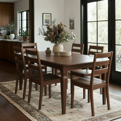 A modern dining room with The Fuller Dining Table featuring extension leaves and six matching chairs on a patterned rug. A vase with dried flowers sits atop the table, framed art decorates the wall, and large windows fill the space with light.