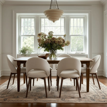A bright dining room showcases The Fuller Dining Table with extension leaves, six white upholstered chairs, a floral centerpiece, a chandelier, tall windows, and a patterned rug covering the wooden floor in this handcrafted USA space.
