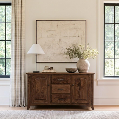 The Gibson Sideboard Table is a handcrafted hardwood piece with two cabinets flanking three central drawers, topped with a lamp, a stack of books, a bowl, and a vase of greenery.