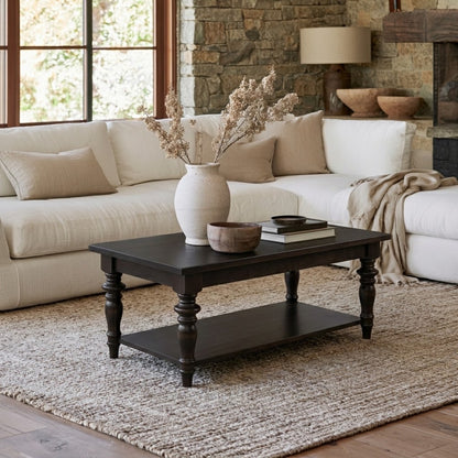 A cozy living room with a white sofa features The Lincoln Rectangle Coffee Table, a dark, handcrafted solid wood piece made by American craftsmen and finished with a durable top coat. Resting on a textured rug and set against a stone wall with a window in the background, the coffee table holds books, bowls, and a vase of dried flowers—adding timeless charm to the inviting space.
