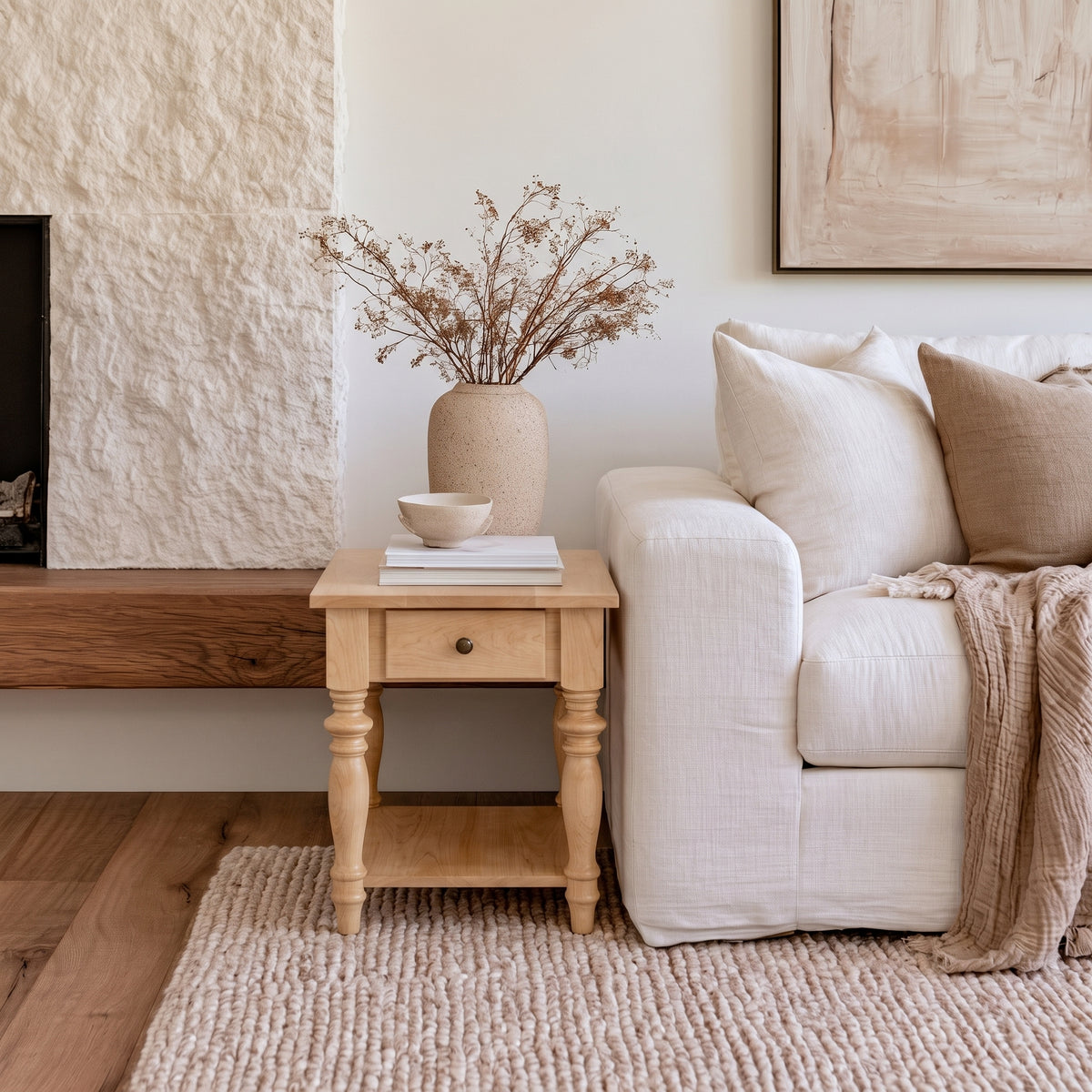 A cozy living room corner features a white sofa, beige pillows, a knit throw, and The Lincoln End Table—a handcrafted solid wood side table displaying a vase of dried flowers and books. A textured rug lies on the wooden floor beside a stone fireplace, evoking timeless charm inspired by American craftsmen.