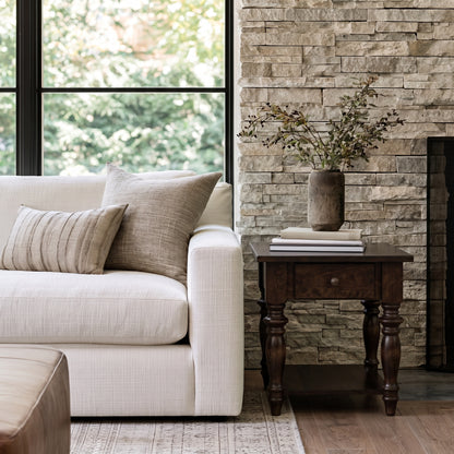 A white sofa with beige pillows sits beside The Lincoln End Table, a handcrafted solid wood piece built by American craftsmen and finished with a durable top coat, holding books and a vase with leafy branches. Behind, a stone wall and large window reveal greenery outside.