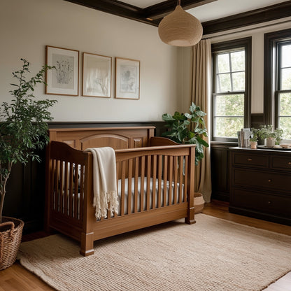 A cozy nursery features The Macon Crib with a white blanket, framed art, a soft rug, potted plants, a dresser, and large windows that fill the space with natural light.