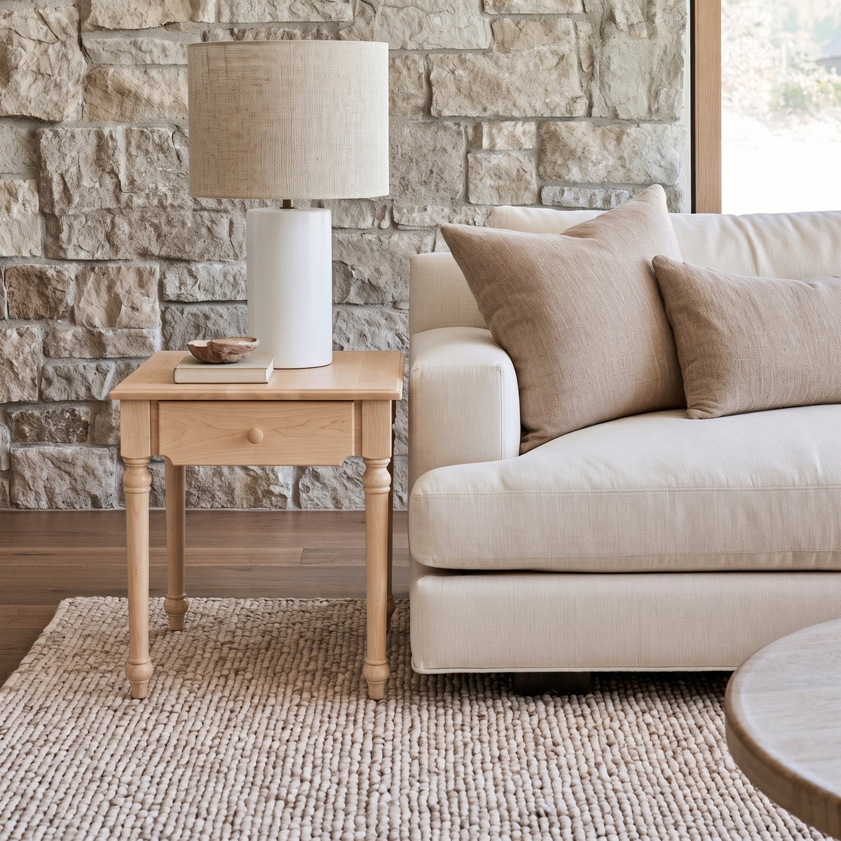 A neutral-toned living room with a beige sofa, matching pillows, The Marcy End Table—a handcrafted solid wood side table made by American craftsmen and finished with a durable top coat—topped with a white lamp and a small dish, a textured rug, and a stone wall in the background.