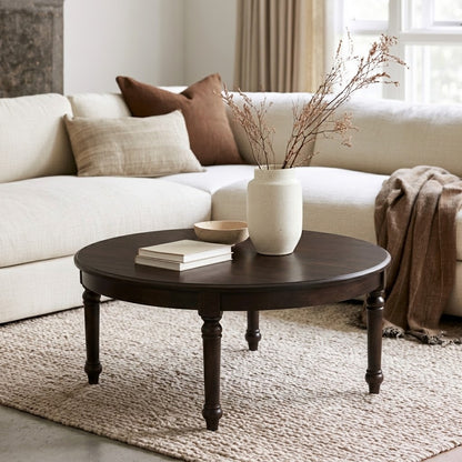 The Marcy Round Coffee Table—a dark, solid wood piece handcrafted in the USA—sits atop a textured rug in front of a cream sectional sofa adorned with neutral pillows and a brown throw. Styled with books, a ceramic vase filled with dried branches, and a bowl, this coffee table’s durable top coat ensures lasting beauty and resilience for everyday living in your bright, cozy living room.
