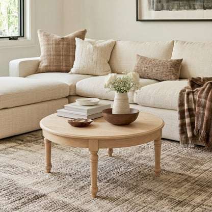 The Marcy Round Coffee Table, handcrafted in the USA from solid wood and finished with a durable top coat, sits in front of a beige sectional sofa adorned with pillows and a plaid throw. Styled with books, a vase of white flowers, and bowls, this timeless piece brings lasting quality to a cozy, sunlit living room.