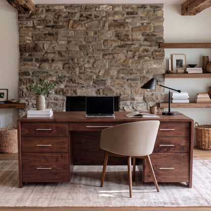 A dark walnut wood writing desk with six drawers and silver handles, styled with a beige upholstered chair, laptop, and lamp in front of a rustic stone wall.