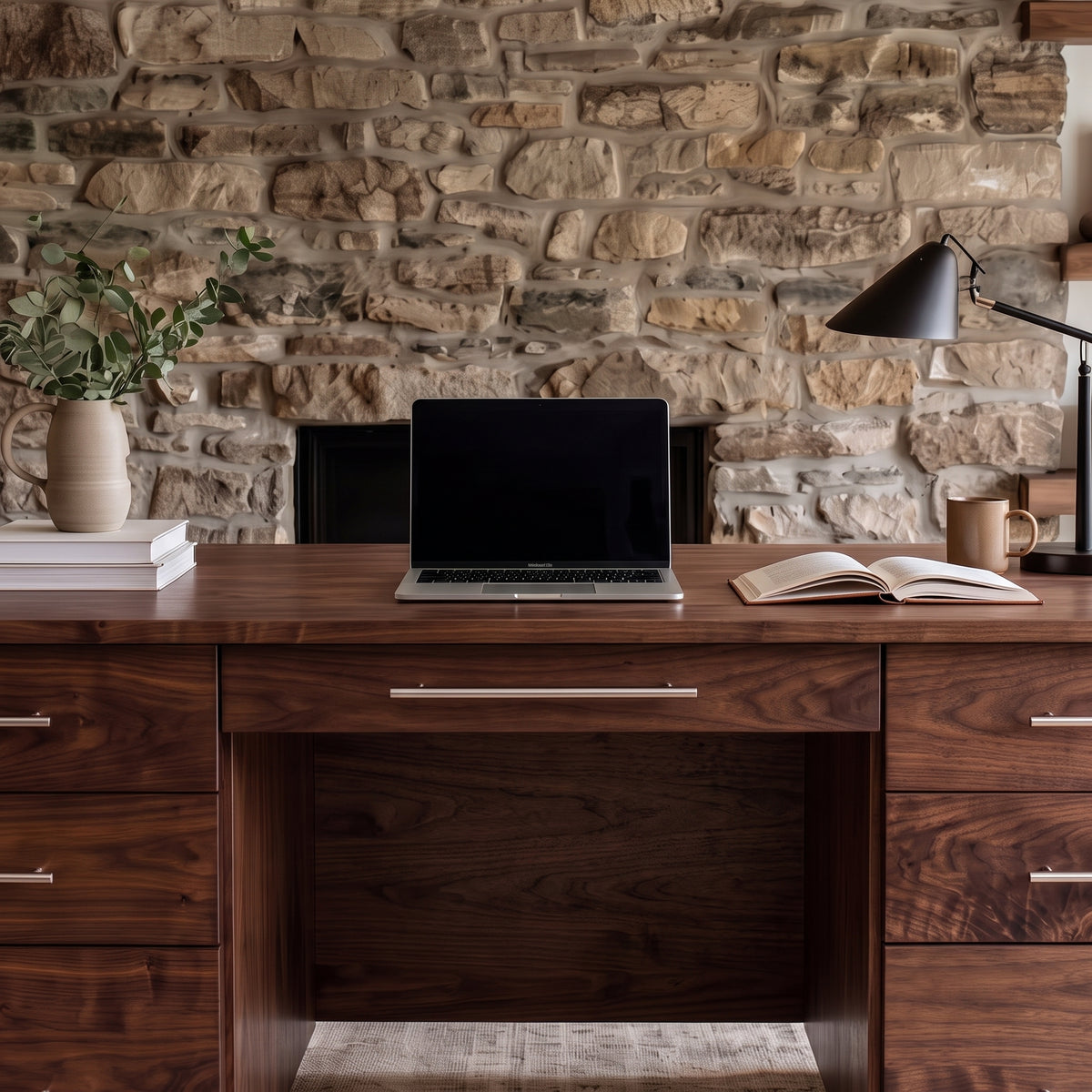 A dark brown wooden writing desk with silver metal drawer pulls, styled with a laptop, books, a lamp, and a mug in front of a rustic stone wall.