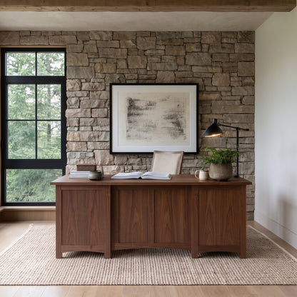 An open book, a potted plant, and a desk lamp sit on The Owen Hardwood Desk, which is placed against a stone wall in front of a window.