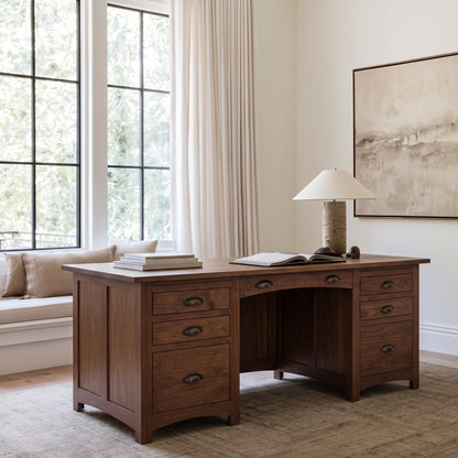 The Owen Hardwood Desk is a handcrafted solid hardwood desk with a row of drawers on each side, topped with a lamp and an open book.