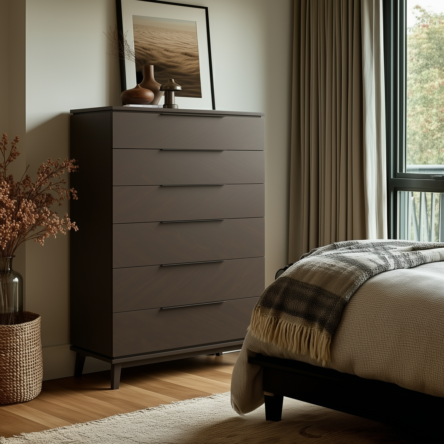 A modern bedroom features The Adelaide Chest of Drawers in dark wood, decorative vases, framed art, and a cozy bed with a plaid blanket. Natural light fills the room, highlighting a woven basket with dried plants on the wooden floor.