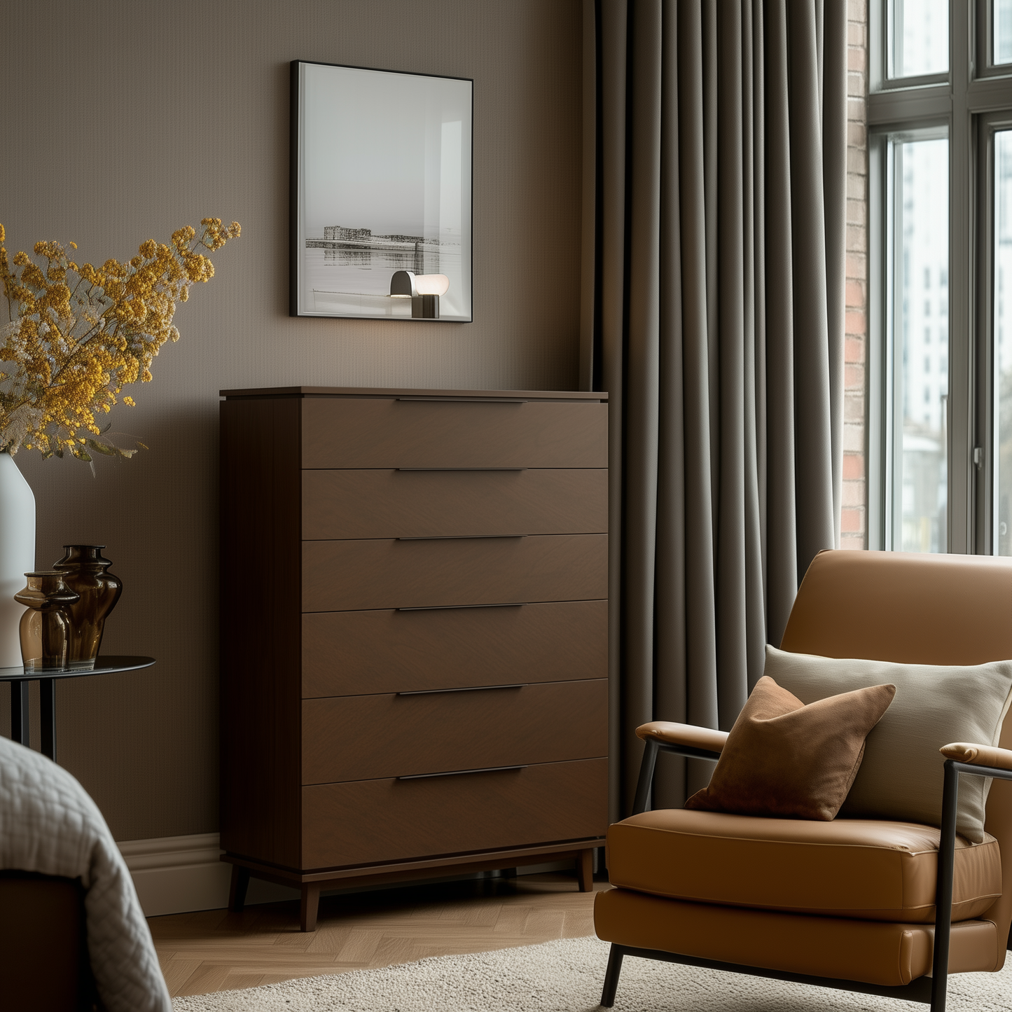 A modern bedroom corner featuring The Adelaide Chest of Drawers, a beige armchair with cushion, a vase of yellow flowers, gray curtains, and a black-and-white photo above the dresser. Sunlight pours in from a nearby window.