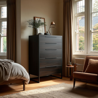 A tidy bedroom corner featuring The Preston Chest of Drawers in dark wood, a small side table with a vase, a brown armchair, and large windows with beige curtains allowing natural light.