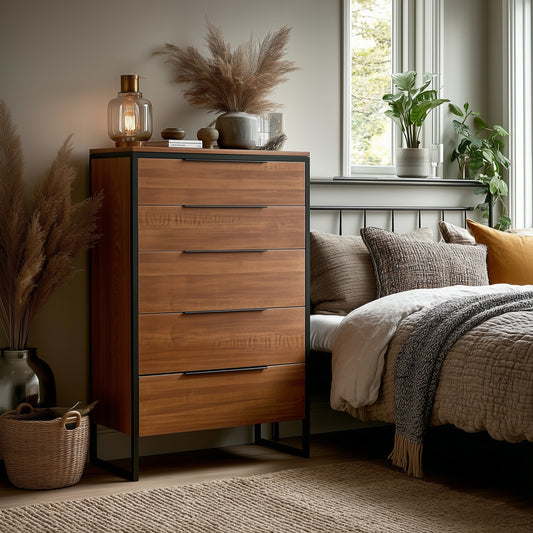 A cozy bedroom showcases The Preston Chest of Drawers, a tall wooden piece with five drawers, decorated with pampas grass, a lamp, and books. The bed is neatly layered with blankets and cushions as sunlight streams through the window.