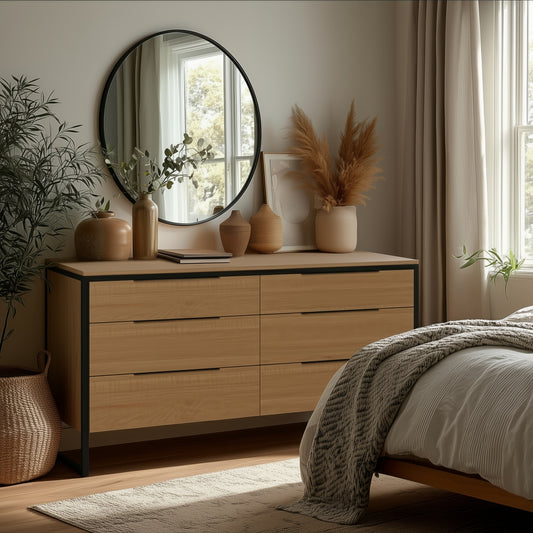 A cozy bedroom corner features The Preston 6-Drawer Dresser topped with vases, pampas grass, and a round mirror. Sunlight streams through beige curtains, highlighting part of a neatly made bed.