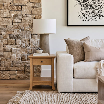 A cozy living room with a white sofa, neutral pillows, The Veris End Table topped with a lamp, a textured rug, a stone accent wall, and framed botanical art—featuring solid wood furniture handcrafted in the USA and finished with a durable top coat for lasting beauty.