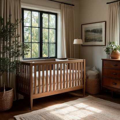 A cozy nursery features The Westcott Crib, beige bedding, a dresser, wicker basket, potted plants, a rug, and a large window with light curtains allowing natural light in. A framed landscape artwork decorates the wall.