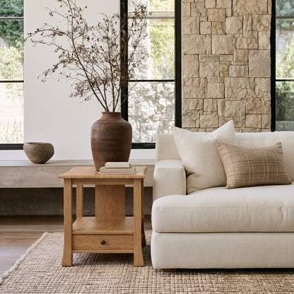 A neutral-toned living room featuring a cream sofa, The Winslow End Table—a handcrafted solid wood piece—displaying books and a large vase with dried branches, complemented by a stone accent wall and expansive windows that fill the space with natural light.